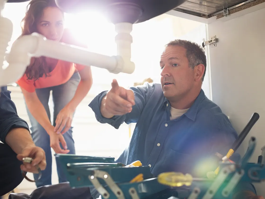 Master plumber explaining a repair to a homeowner under a kitchen sink